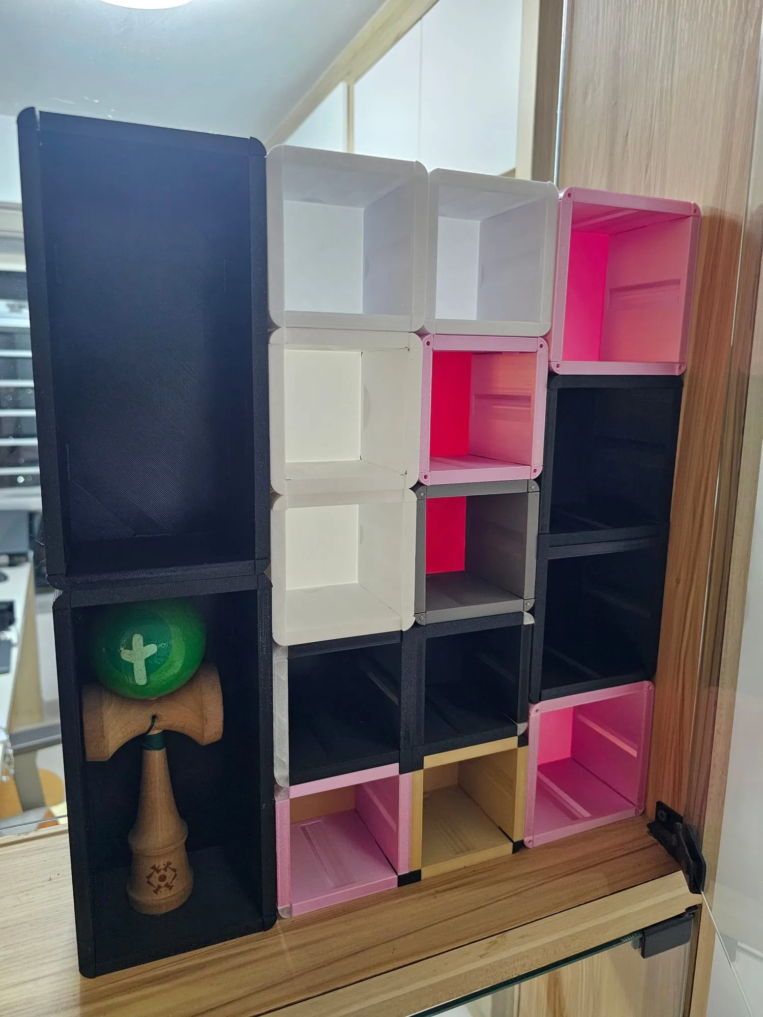 Two black ThrowGRID kendama cubes forming the left-hand column of a mixed wall, the lower one holding a green and wood kendama, with columns of 1A cubes alongside